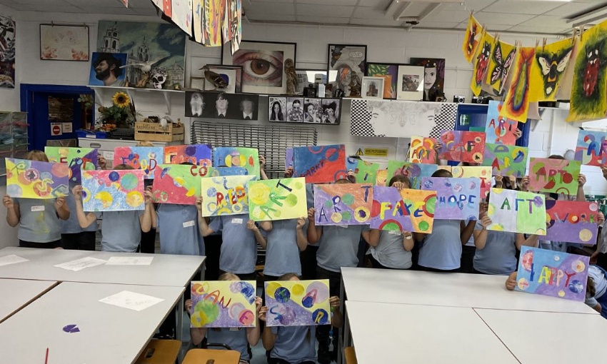 A group of primary school pupils in an art classroom holding their newly created colourful artworks in front of their faces